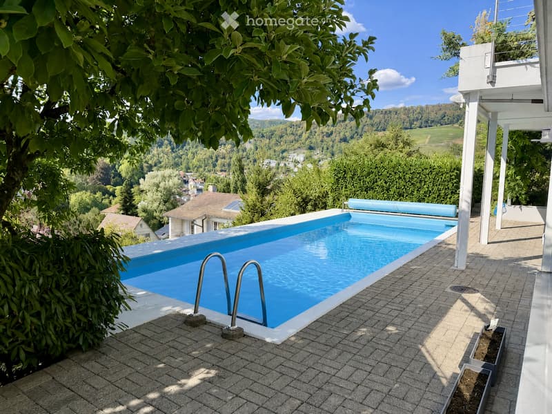 Outdoor area with a rectangular swimming pool surrounded by a tiled deck, featuring a scenic view of a valley and trees.