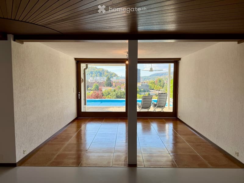 A spacious hallway with tiled flooring, large windows offering a view of a pool and surrounding landscape, and a central column separating the space.