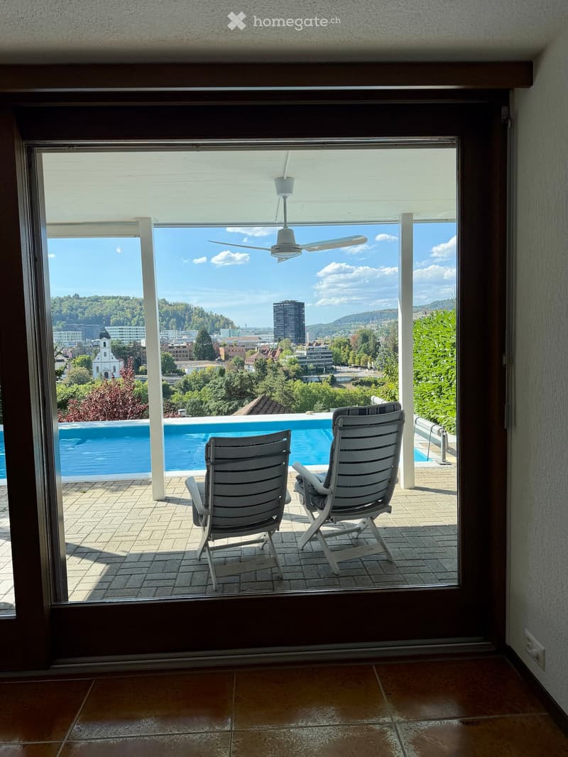 Balcony with two white chairs, overlooking a swimming pool and a cityscape with trees and buildings.