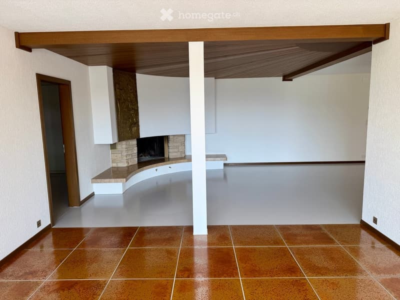 Empty room with tiled floor, white walls, and a fireplace with a wooden mantle. There is a wooden ceiling and a doorway leading to another room.