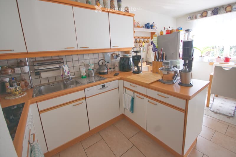 Bright kitchen with white cabinets, wooden trim, and tiled backsplash. Features a sink, dishwasher, oven, refrigerator, coffee maker, mixer, and various kitchen utensils on the counter.