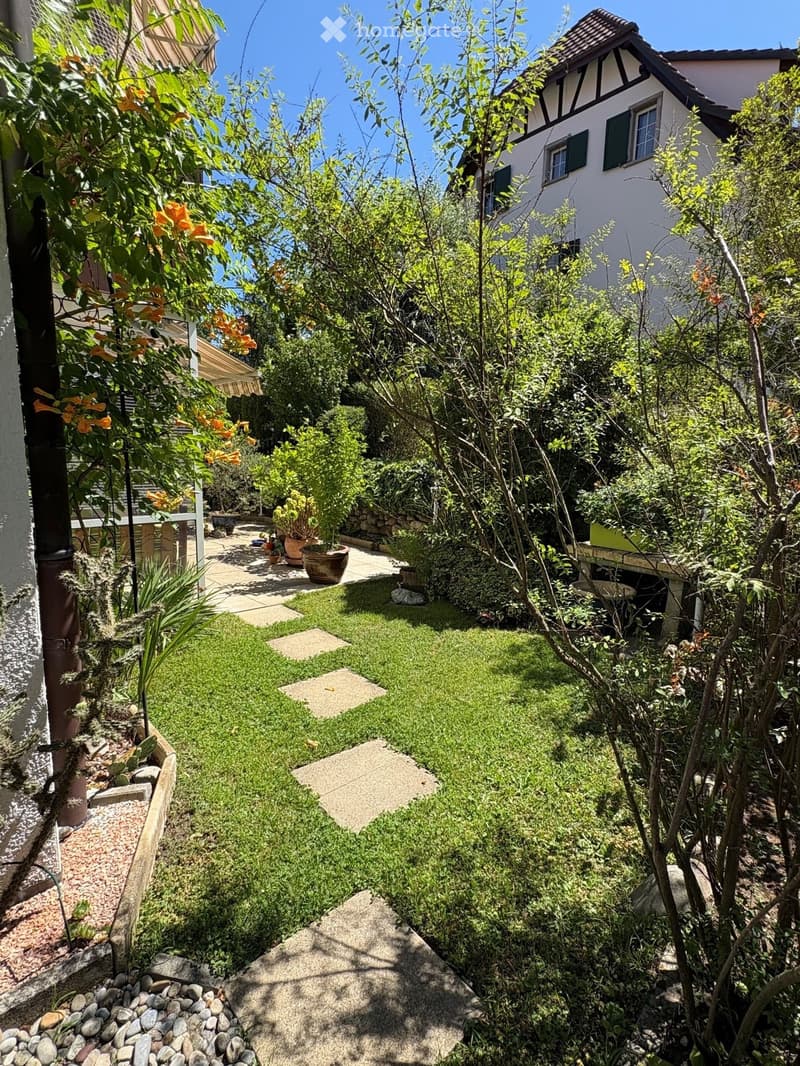 A well-maintained garden with a mix of green grass, shrubs, trees, and potted plants. The garden features a stone pathway and a small seating area with a table and chairs.