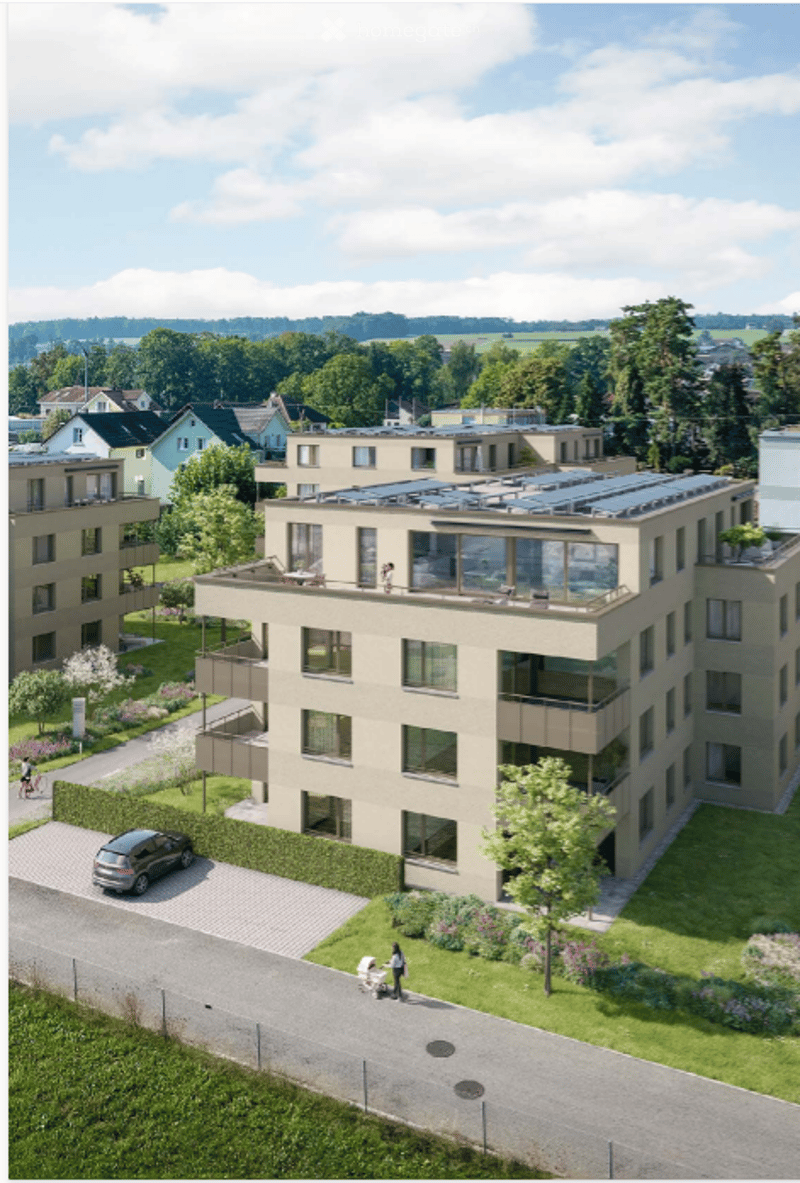 A modern multi-story residential building with balconies, large windows, and solar panels on the roof. The surrounding area includes greenery, a driveway, and neighboring houses.