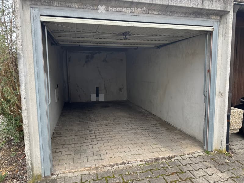Garage with concrete walls, brick floor, and an open garage door leading to an outdoor area.