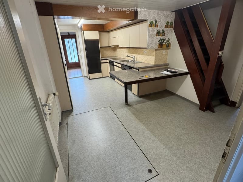 Hallway leading to a kitchen area with cabinets, sink, and countertop. Light-colored walls, tiled floor, and a radiator on the left side.
