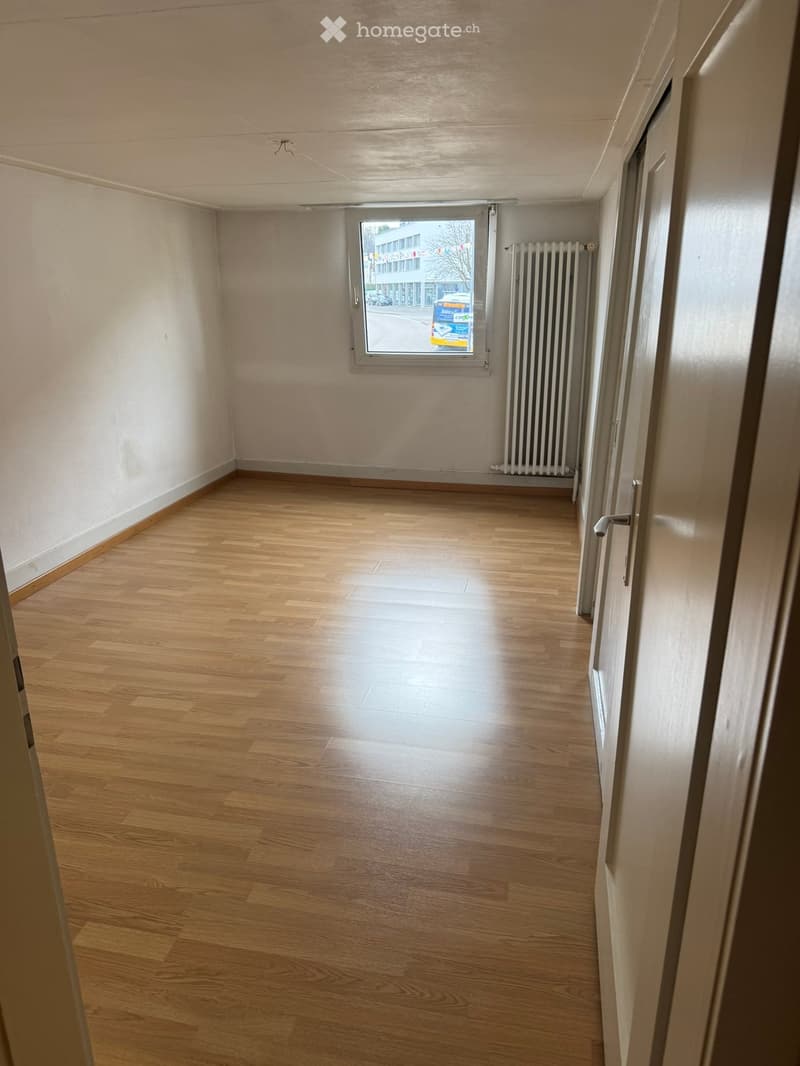Empty room with wooden flooring, white walls, a window with a view of the street, and a radiator on the wall.