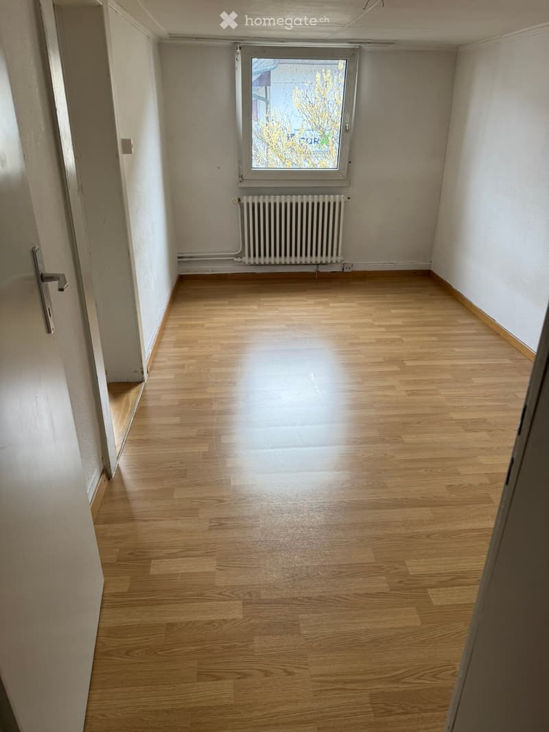 Empty hallway with wooden flooring, white walls, a radiator, and a window showing a view of trees outside.