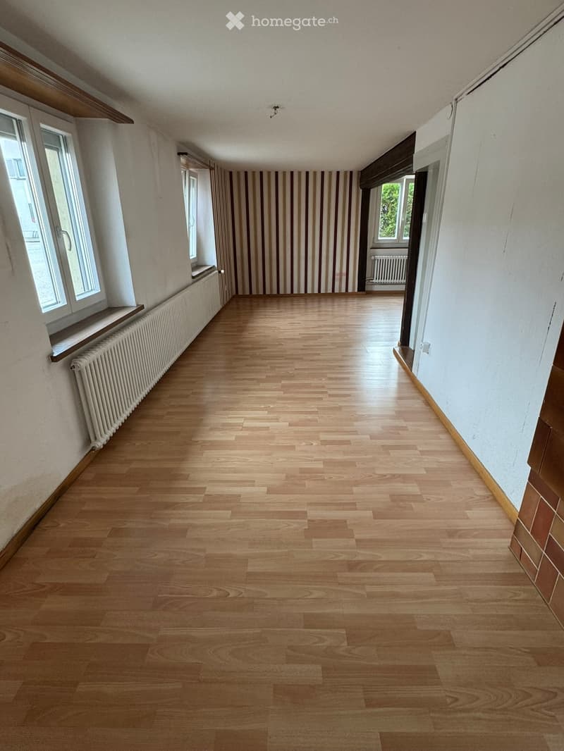 Empty hallway with wooden flooring, white walls, and large windows on one side. Radiators are visible along the walls.