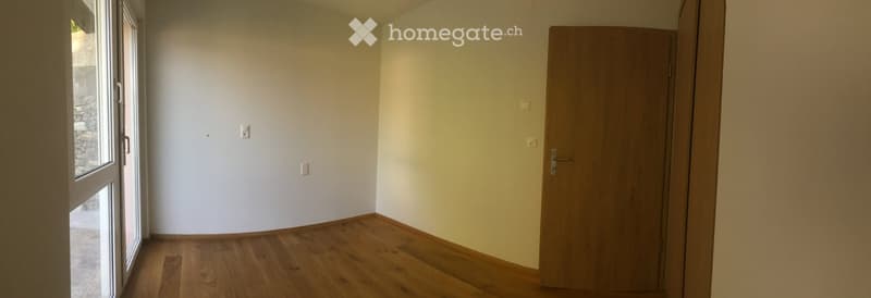 Empty room with wooden flooring, white walls, and a large window with a view of a stone wall outside. A closed wooden door is visible on the right side.