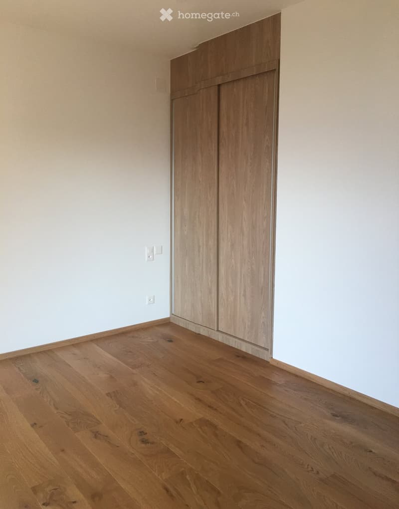 Empty room with light wooden flooring and white walls, featuring a built-in wooden closet with double doors.
