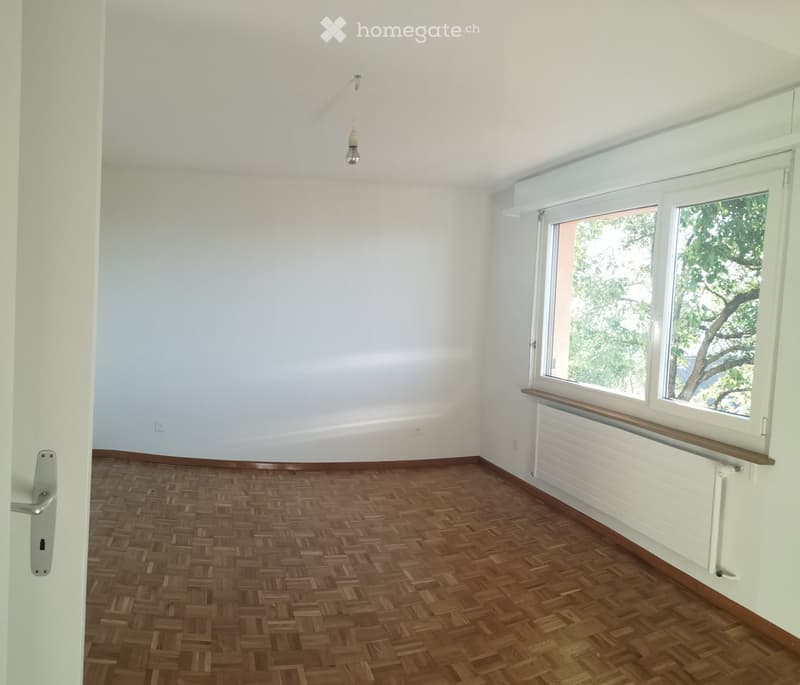 Empty room with wooden herringbone flooring, white walls, large windows with wooden sills, and a radiator below the windows. A single light fixture hangs from the ceiling.