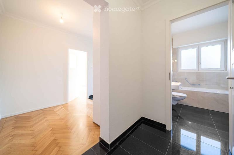 Bathroom with a bathtub, toilet, and sink. The hallway has wooden flooring and white walls.