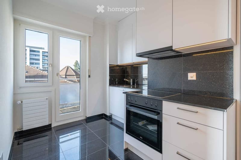 Modern kitchen with white cabinets, black countertops, and a built-in oven. Features a large window and a sliding glass door leading to a balcony.