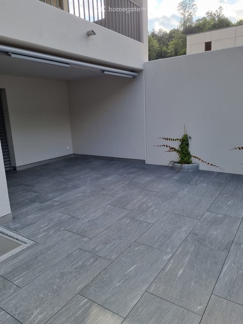 Grey tiled balcony with a small potted plant and a modern white building in the background.