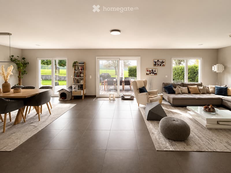 Open-plan living and dining area with modern furniture, large windows, and a view of a garden. Features a sectional sofa, coffee table, ottoman, dining table with chairs, and decorative items.
