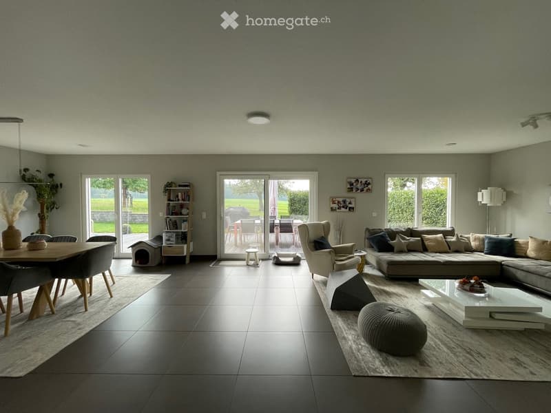 Modern living and dining area with a grey sectional sofa, white coffee table, dining table with chairs, and a pet bed in the corner. Large windows and sliding doors lead to an outdoor patio.