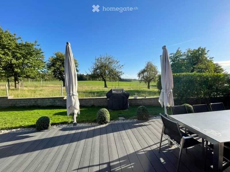 A spacious outdoor patio with wooden decking, surrounded by a well-maintained lawn and trees. Features include a dining table with chairs, a grill, and two large umbrellas.