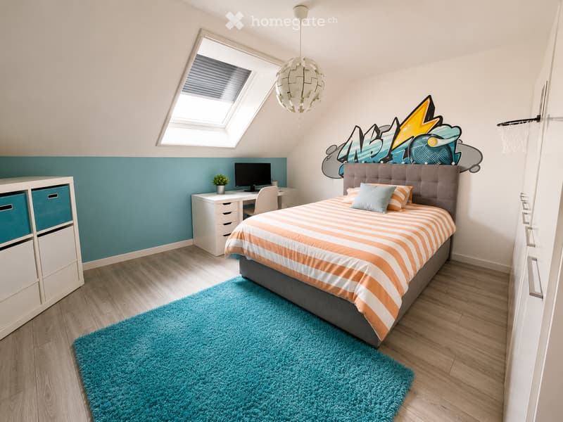 Modern bedroom with a large bed, striped bedding, a blue shag rug, a white desk with a computer, and a colorful graphic wall art. The room features wooden flooring and a skylight window.