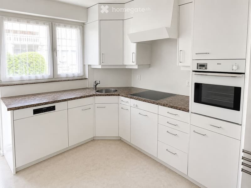 Modern kitchen with white cabinets, granite countertops, stainless steel sink, dishwasher, oven, and range hood. Large windows with white curtains provide natural light.