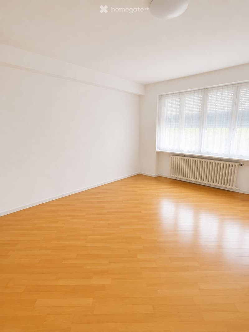 Empty room with light wooden flooring, white walls, large windows with blinds, and a radiator under the windows.