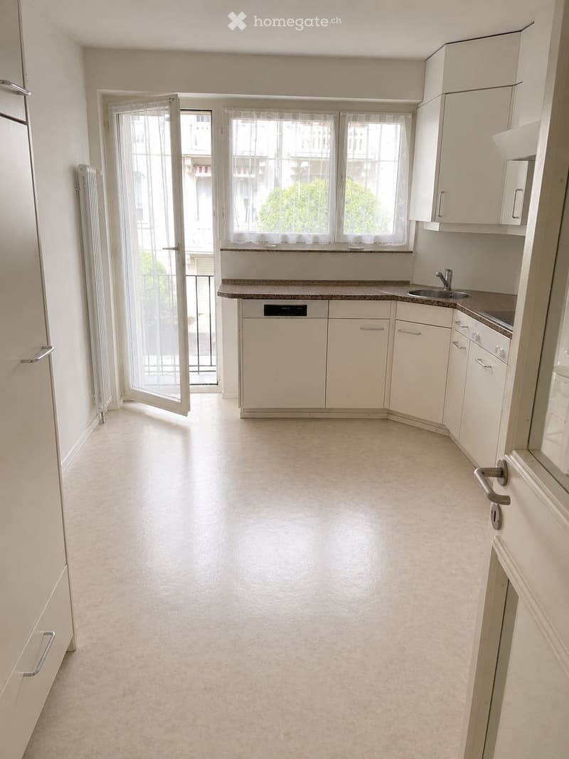 Empty kitchen with white cabinets, a sink, and a dishwasher, featuring a large window and a sliding glass door leading to a balcony.