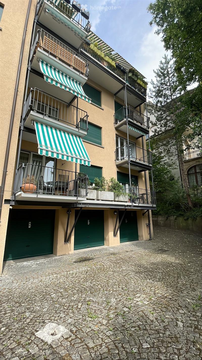 Multi-story residential building with balconies, green awnings, and potted plants. The ground floor has two garage doors, and the balconies feature outdoor furniture and greenery.