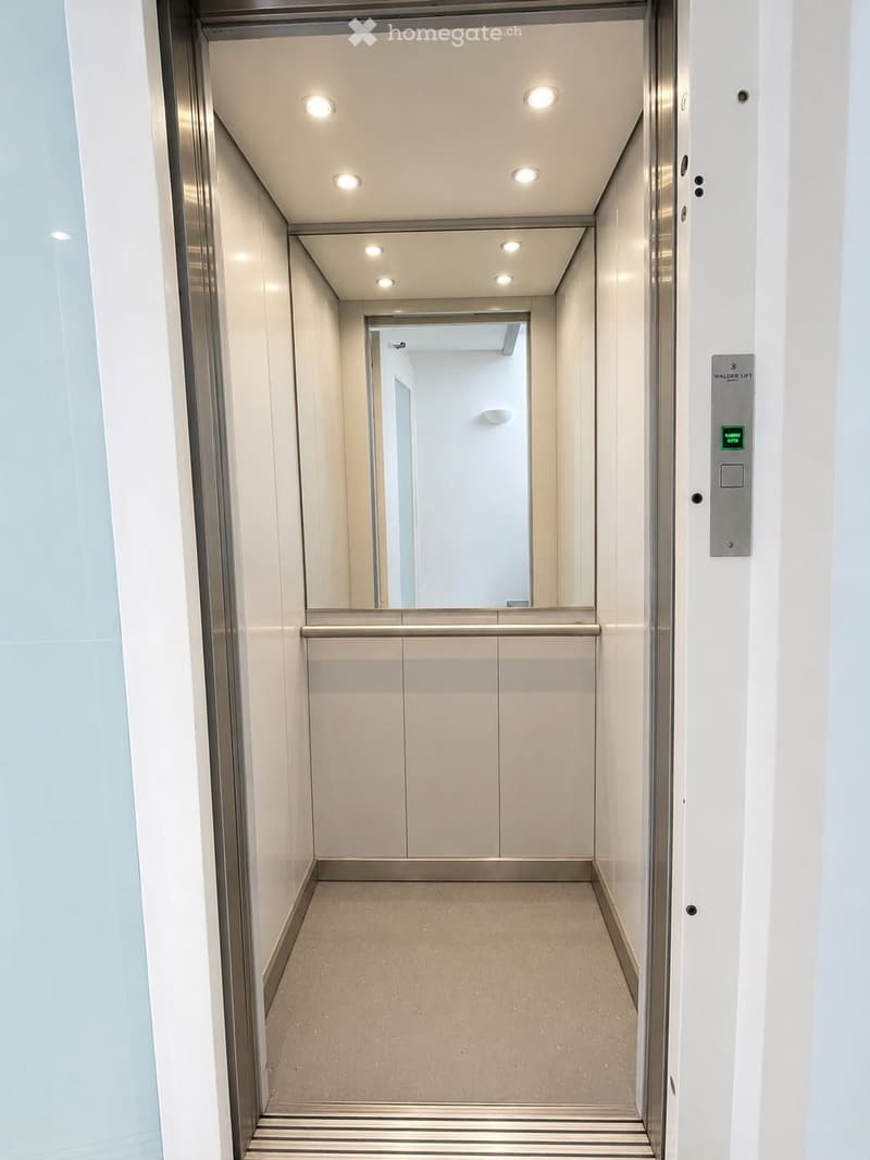 Modern elevator interior with white walls, recessed lighting, and a polished floor. Features a control panel on the right side and a reflective surface at the end.