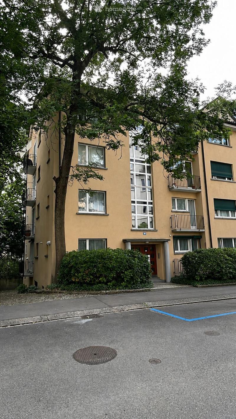 Four-story residential building with beige facade, multiple windows, balconies, and a tree in front.