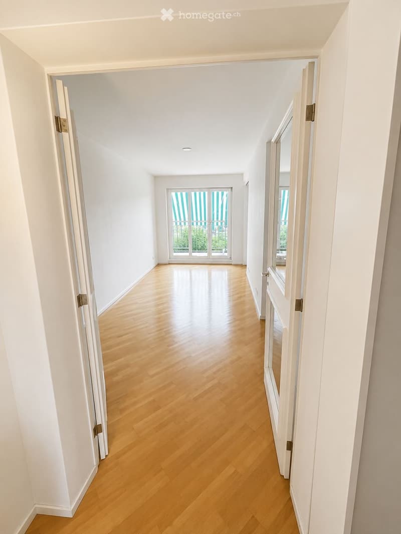 Empty hallway with light wooden flooring, white walls, and recessed ceiling lighting. Features an open doorway leading to a room with large windows and striped blinds.
