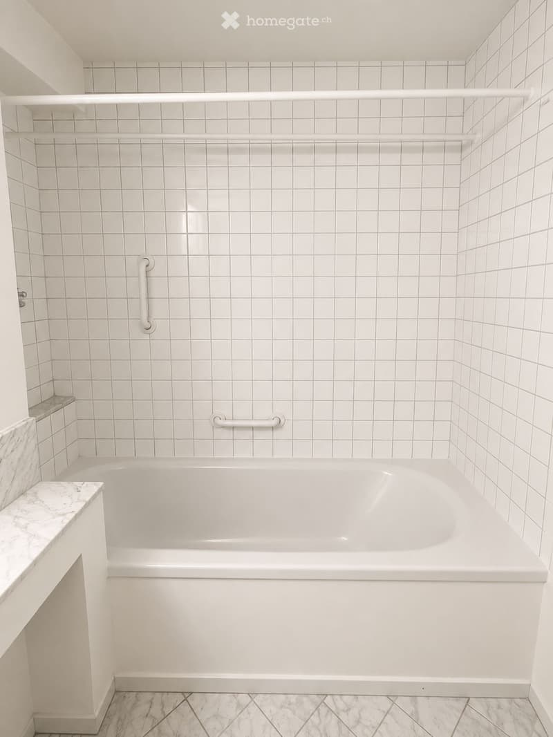 Modern bathroom with a white bathtub, white tiled walls, marble flooring, and grab bars for safety.