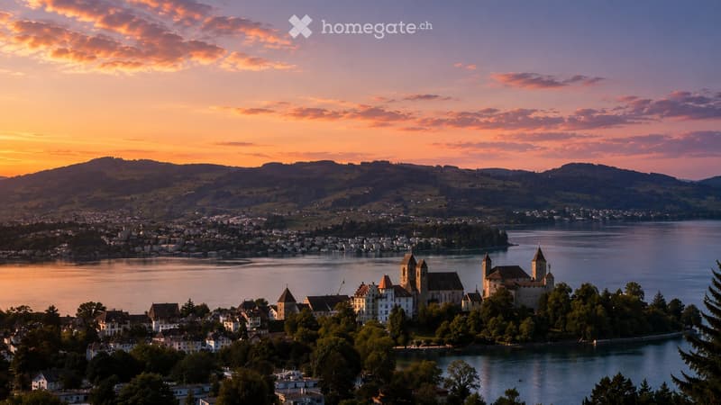 A scenic view of a town situated on a peninsula surrounded by a lake, with historic buildings and a castle-like structure, under a colorful sunset sky.
