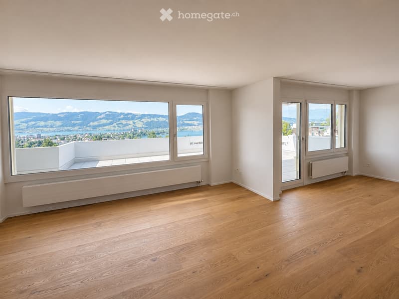Empty room with wooden flooring, large windows offering scenic mountain and lake views, radiators, and a sliding glass door leading to a balcony.