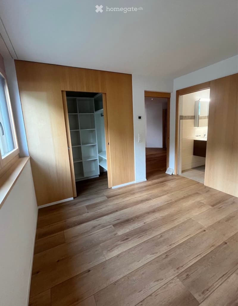 Empty room with wooden flooring, white walls, and built-in shelving unit. Adjacent bathroom visible through open doorway.