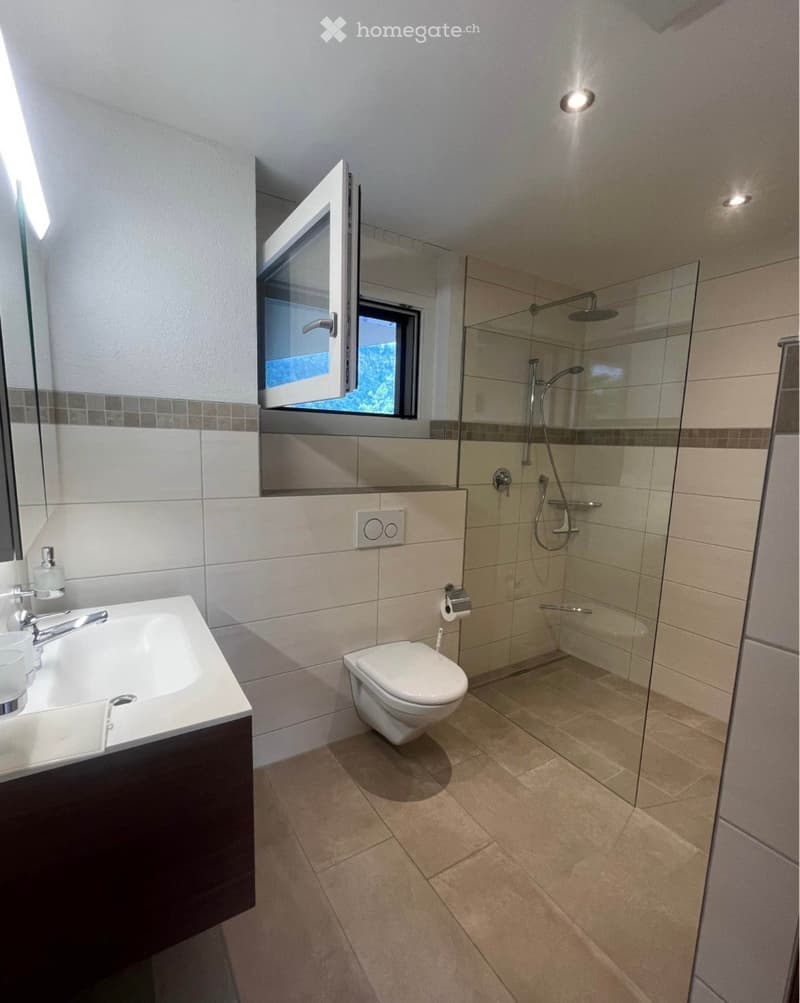 Modern bathroom with a white sink, toilet, glass-enclosed shower, and a window above the toilet. The walls are tiled in beige and grey, and the floor is tiled in light brown.