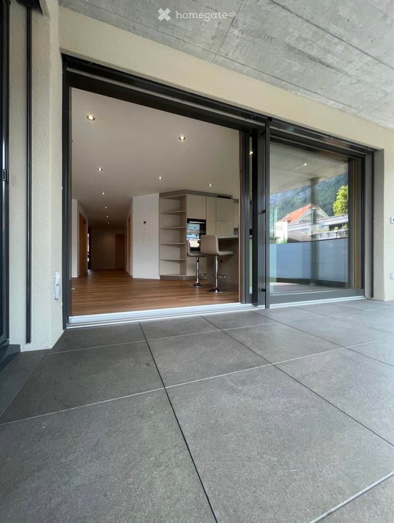 Modern balcony with large sliding glass doors leading to a kitchen area, featuring wooden flooring, white cabinets, and bar stools.