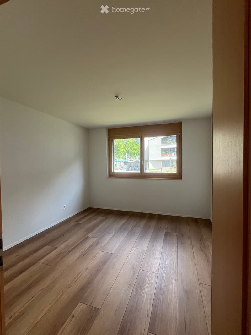 Empty room with light-colored walls and wooden flooring, featuring a large window with a view of the outside.