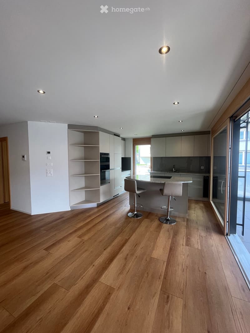 Modern kitchen with light-colored cabinets, a central island with a countertop, two bar stools, and built-in appliances including an oven and sink.