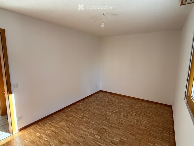 Empty room with wooden flooring and white walls, featuring a single light fixture on the ceiling and a partially open door on the left side.