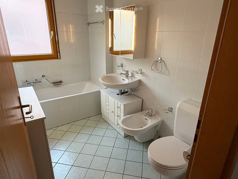 Modern bathroom with a bathtub, sink, toilet, and a mirror above the sink. The room features tiled walls and floor, and a window with wooden frame.
