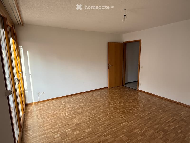 Empty room with wooden flooring, white walls, and large windows allowing natural light. There is a single door leading to another room.