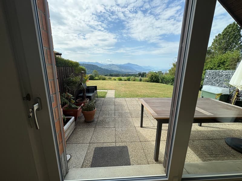 Balcony with tiled floor, wooden table and chairs, potted plants, and a view of a green lawn and distant mountains.