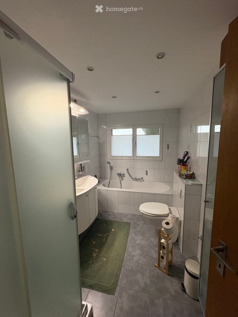 Modern bathroom with a bathtub, toilet, sink, mirror, and a small cabinet. The room features grey tiles, a green bath mat, and a window with blinds.