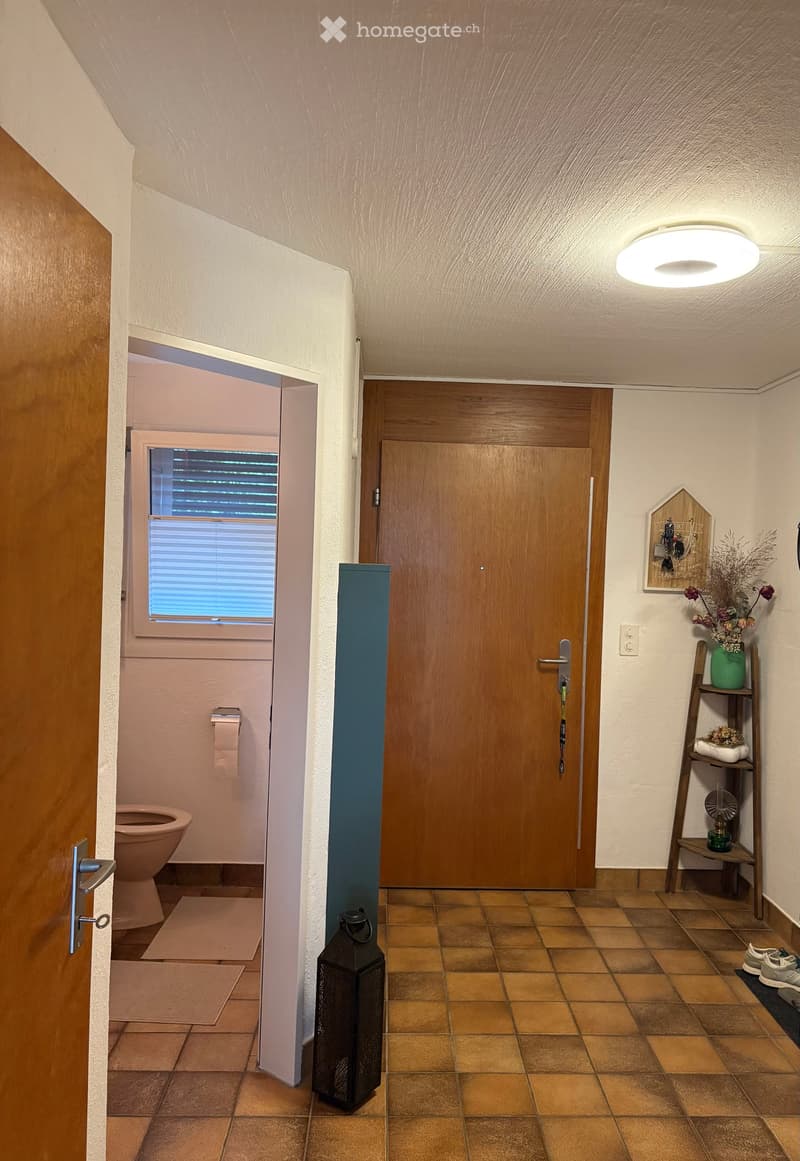 Hallway with tiled floor, wooden doors, a small shelf with decorative items, and a lantern. A toilet is visible through an open door on the left.