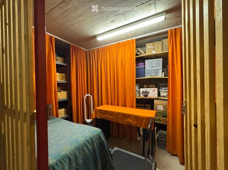 A small, cozy room with wooden walls and ceiling, featuring a bed with a blue blanket, a small table with an orange tablecloth, and shelves with various items behind orange curtains.