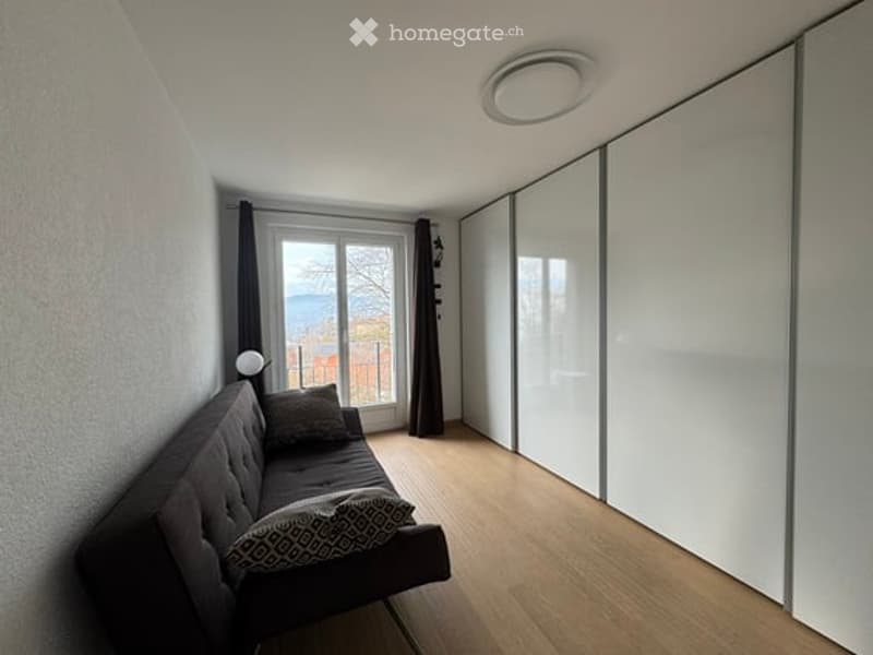 Modern living room with a dark grey sofa, decorative pillows, a round mirror on the wall, and a large sliding wardrobe. The room features wooden flooring and large windows offering a scenic view.
