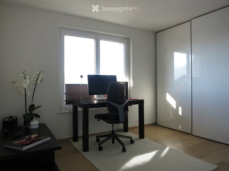 A modern, well-lit room with a desk, office chair, computer, and a small table with a flower and book. The room features large windows and a sleek, minimalist design.