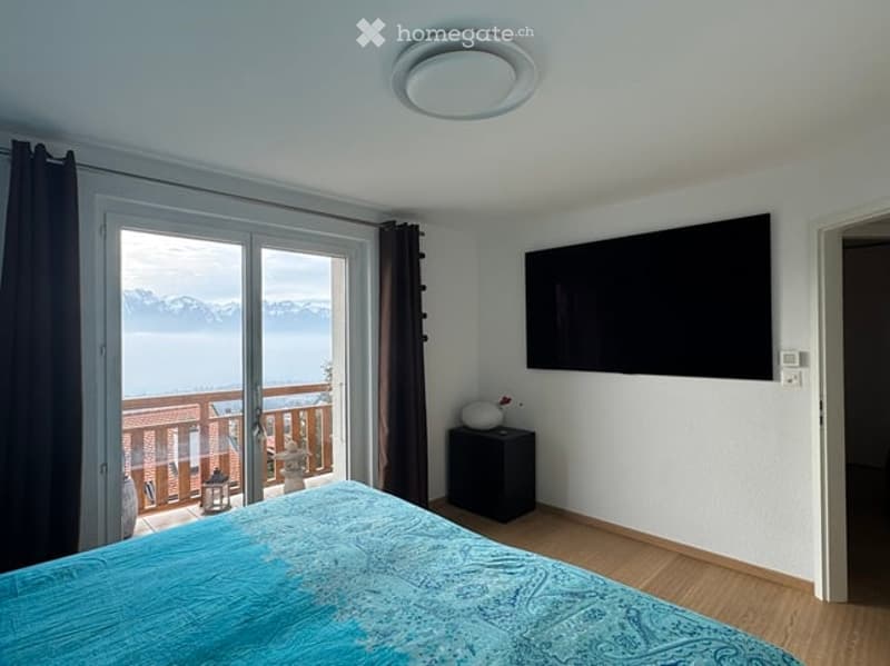 Bright bedroom with wooden flooring, large window offering a scenic view, a bed with a blue duvet, a nightstand with a lamp, and a wall-mounted TV.