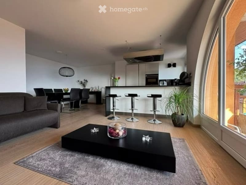 Modern open-plan living and dining area with a grey sofa, black coffee table, dining table with chairs, and a kitchen island with stools. The space features wooden flooring, large windows, and contemporary decor including a plant and flower vase.