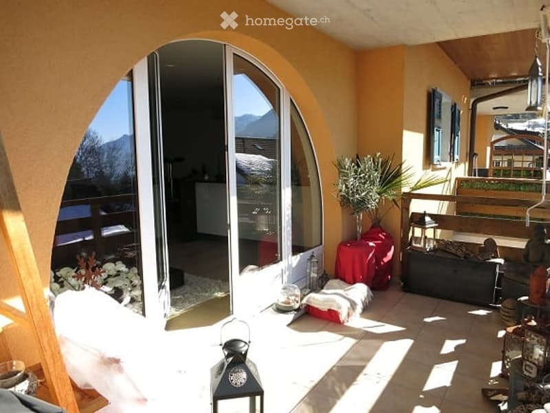 Bright, sunlit balcony with large arched glass doors opening to an indoor space, decorated with cushions, a small table, and potted plants, offering a scenic mountain view.
