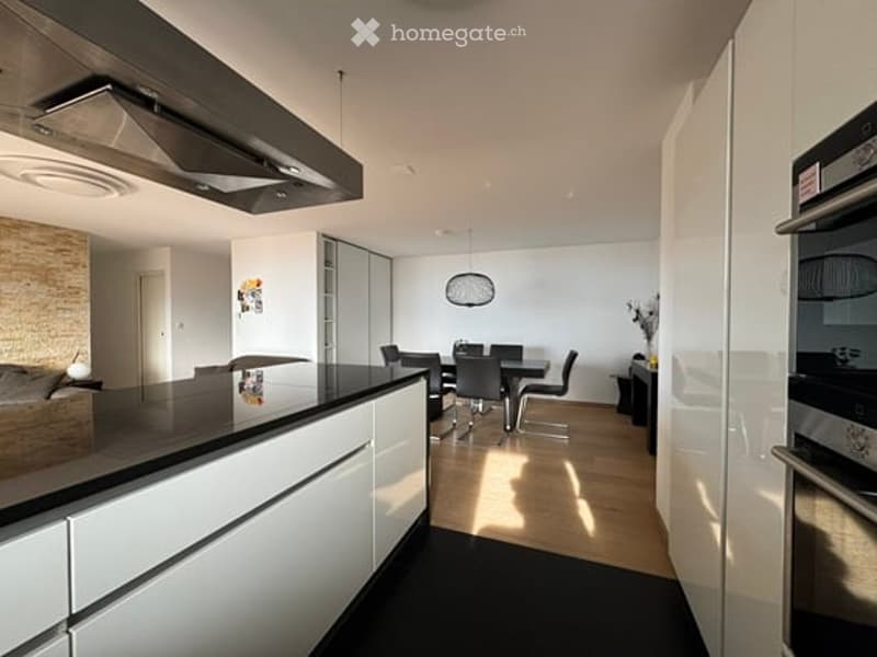 Modern kitchen and dining area with white cabinets, black countertops, a central island, and a dining table with chairs. Features include a built-in oven, refrigerator, pendant light, and a small flower vase on a side table.